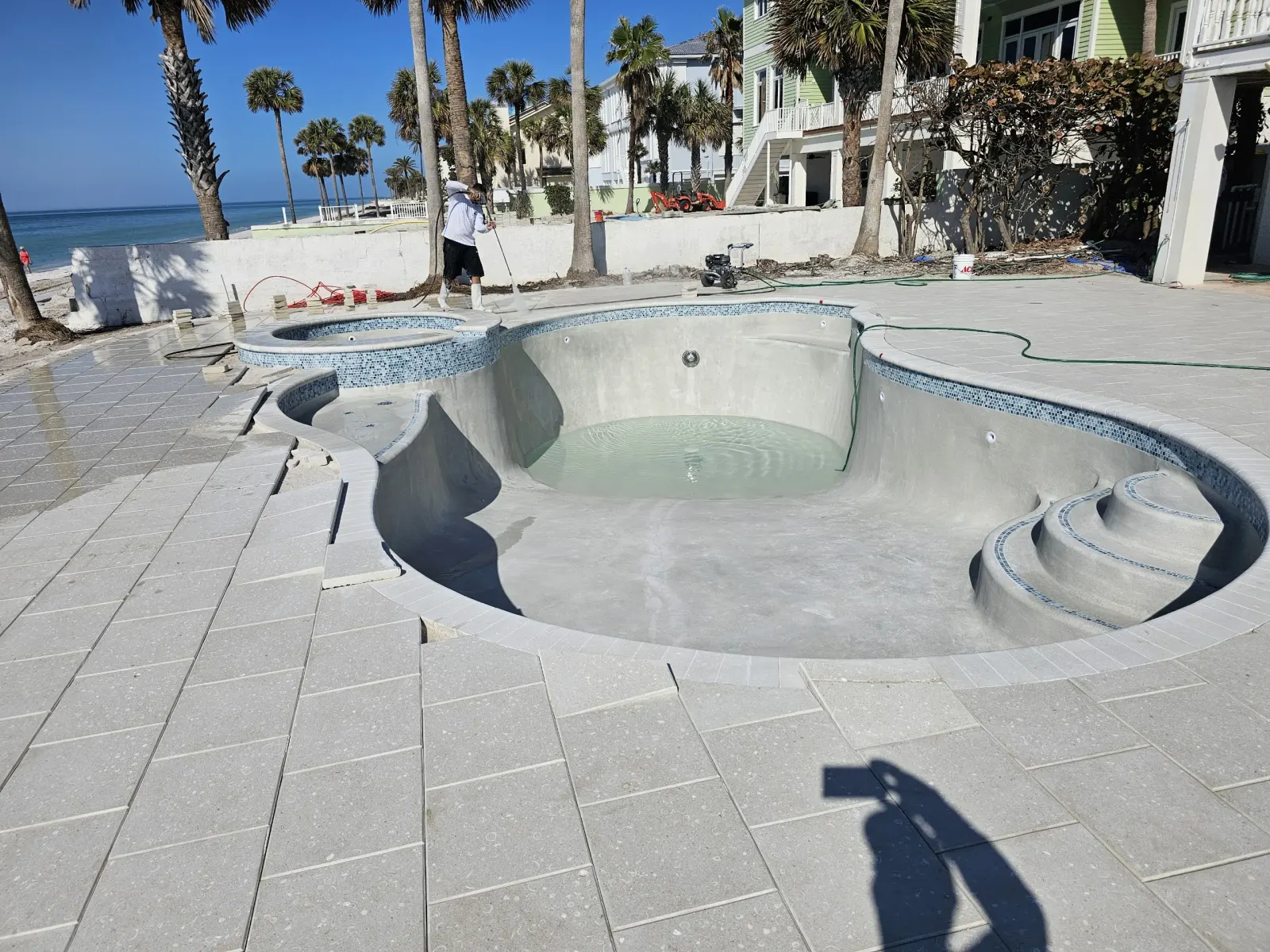 Freeform gunite pool shell mid-construction on a beachfront lot, plaster not yet applied, houses in the background.